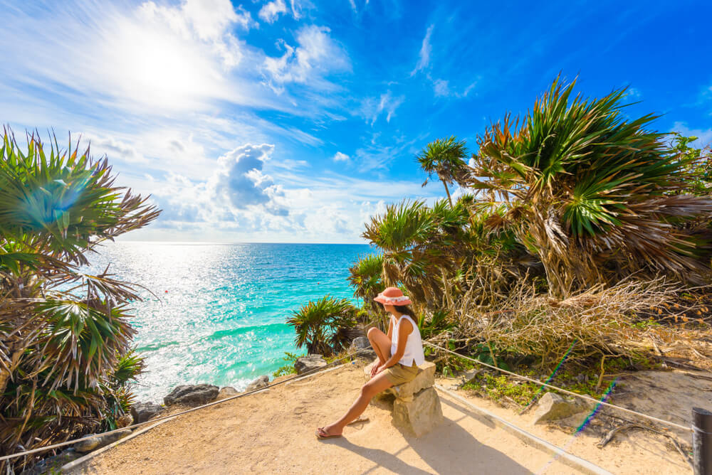A visitor resting on a stone above the turquoise Caribbean, on a path lined with palms