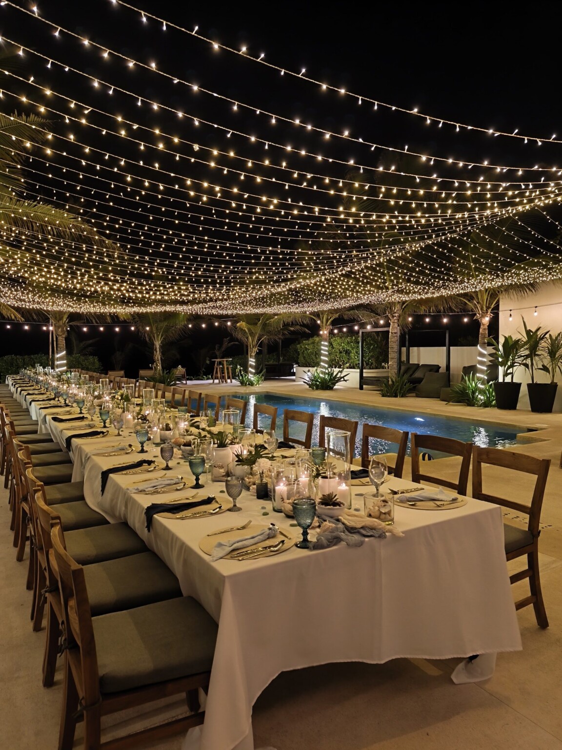 A candlelit long table under a canopy of string lights beside a hacienda pool