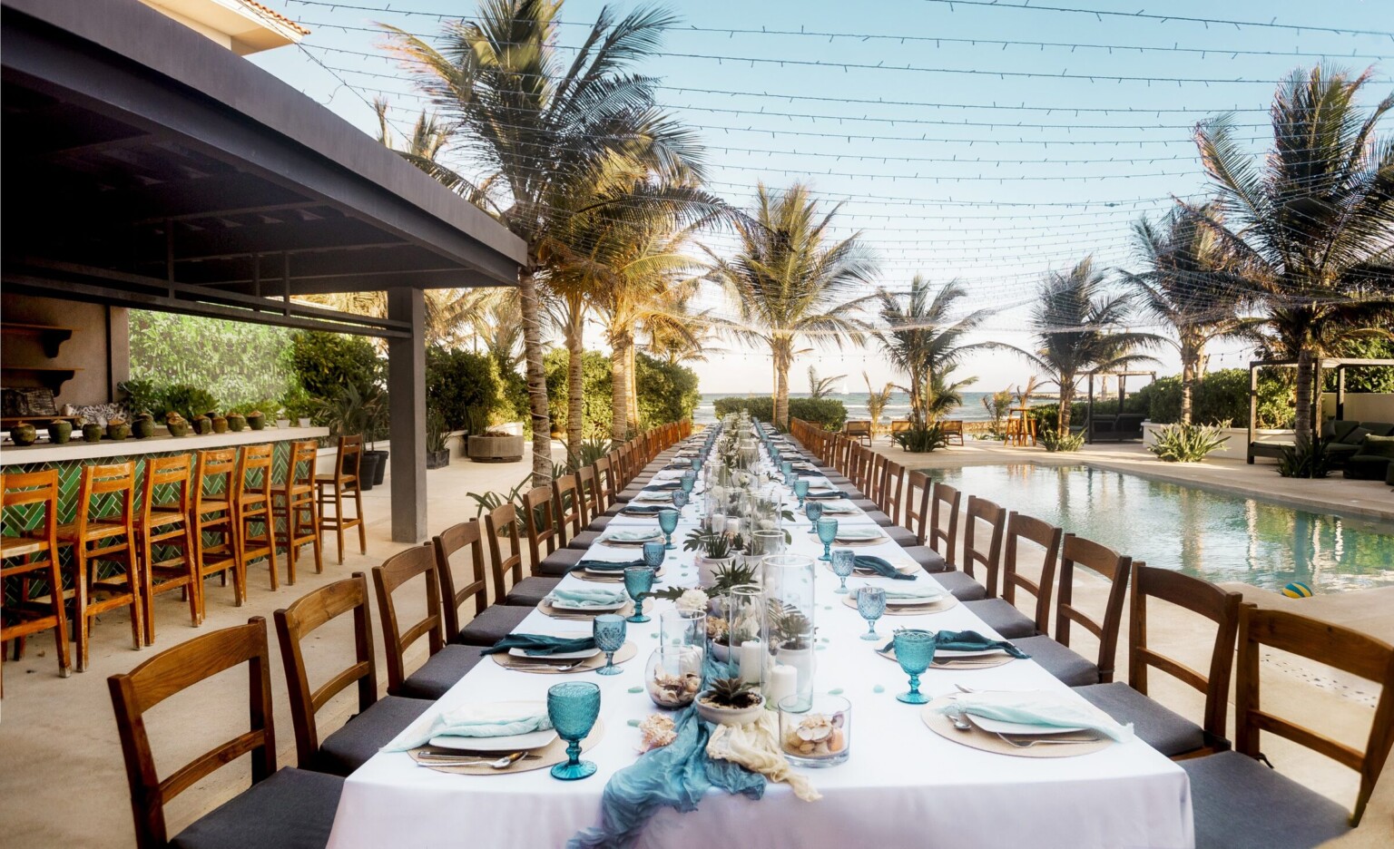 A long dinner table under string lights between the pool and the bar, palms and the sea beyond