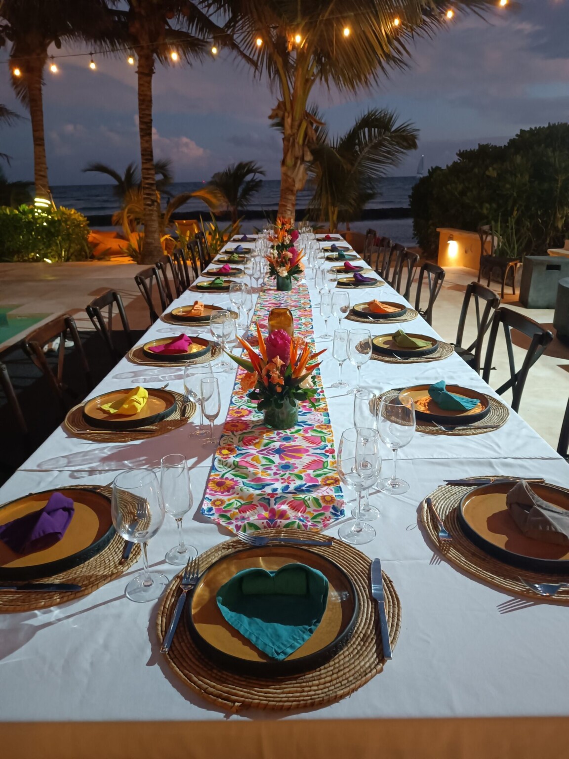 A long table with an embroidered runner and tropical flowers at dusk, palms and the sea behind