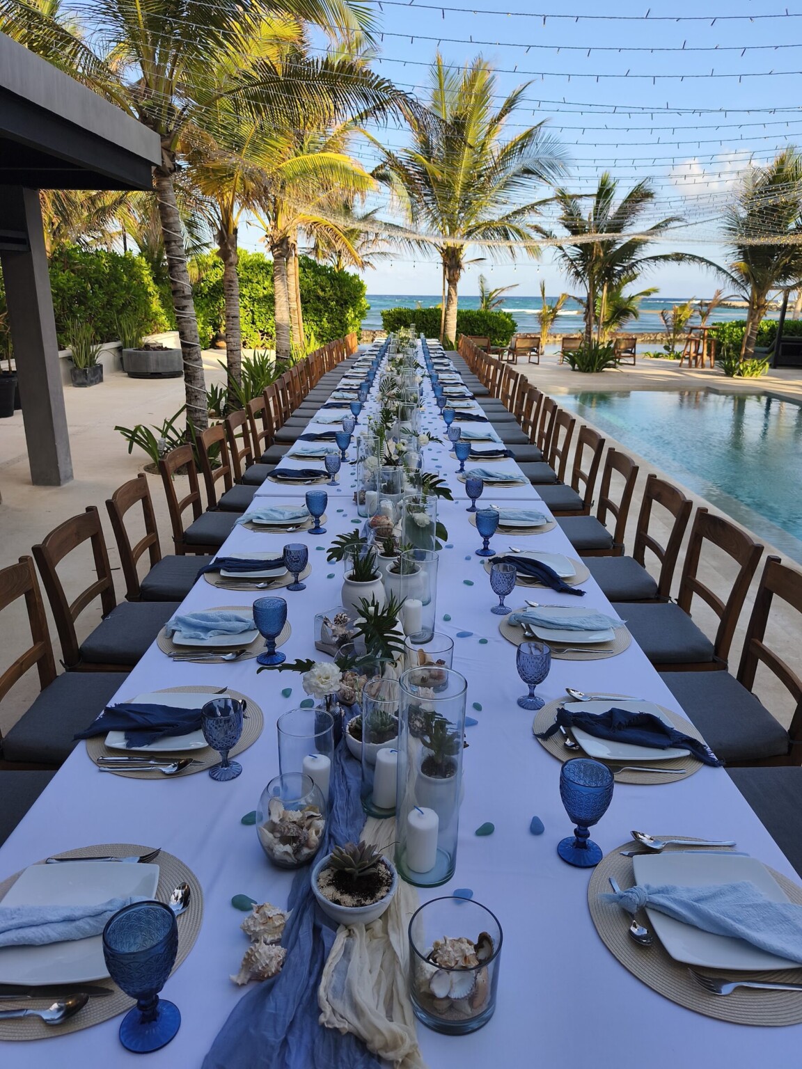 A long dinner table with blue glasses beside a pool that runs to the sea