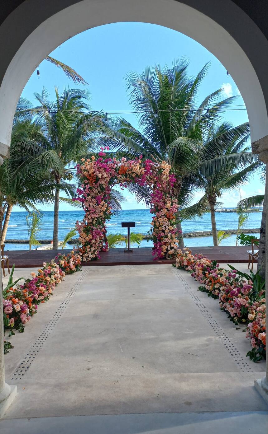 A flower-covered wedding arch framed by a stone arch, the Caribbean behind, at Hacienda Del Mar