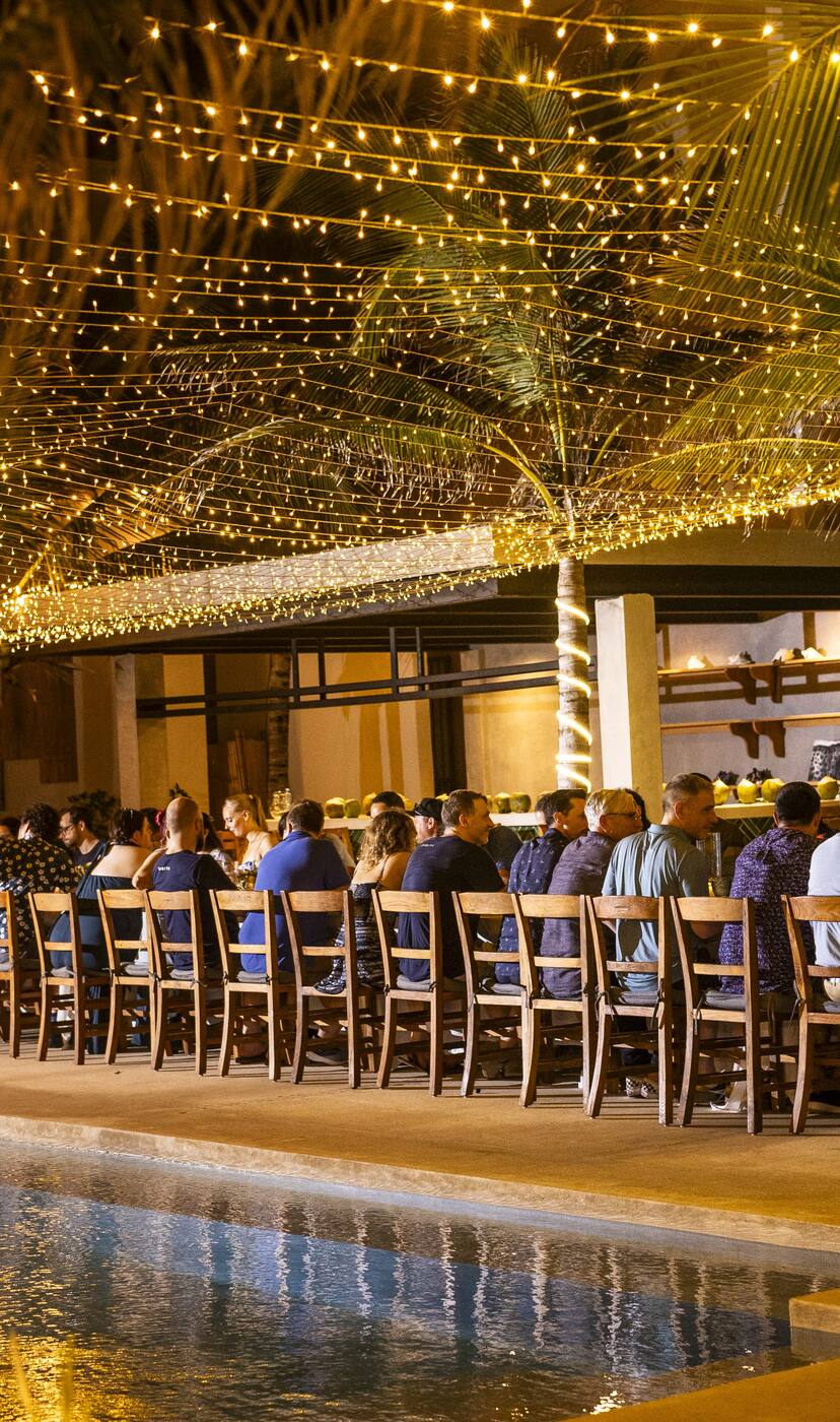 A company dinner at one long table under string lights, beside a hacienda pool