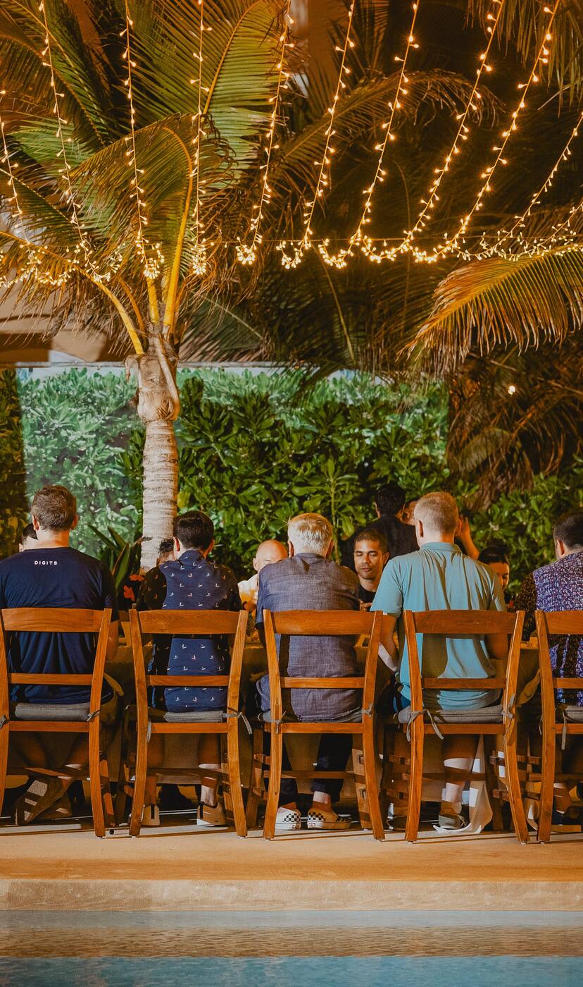 A group at one long table under string lights, beside a hacienda pool at night